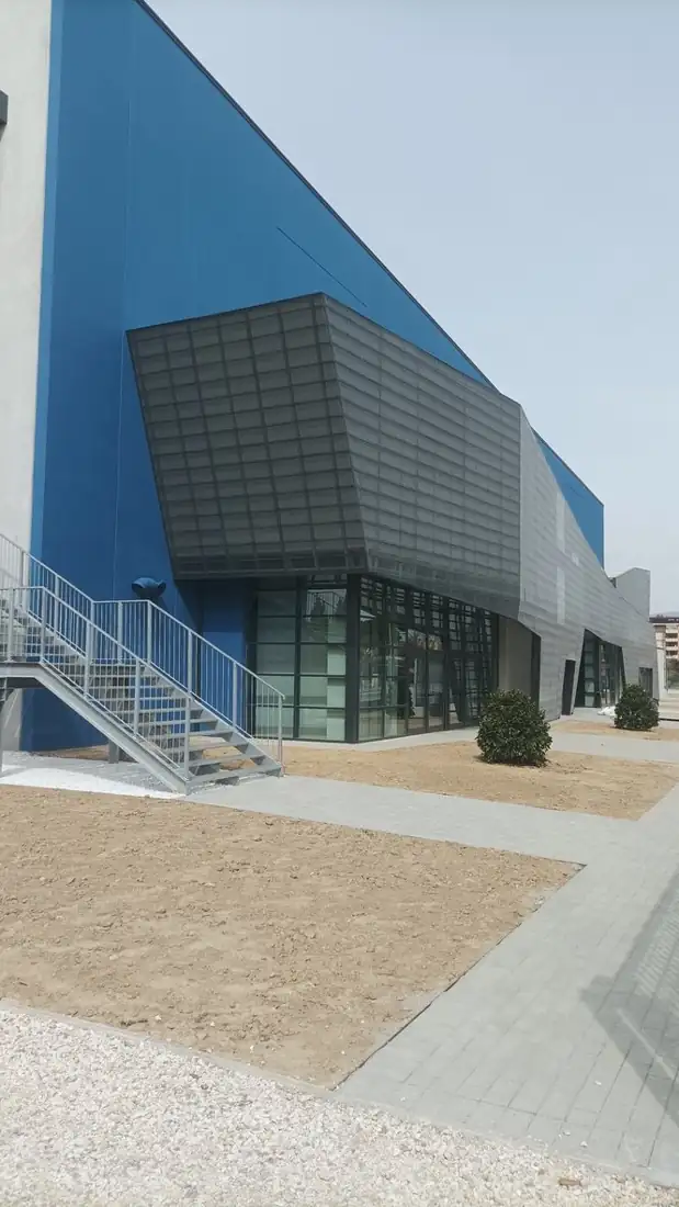 Esterno del palasport: volume blu, rivestimento metallico sfaccettato sopra l'ingresso vetrato e scala esterna in acciaio