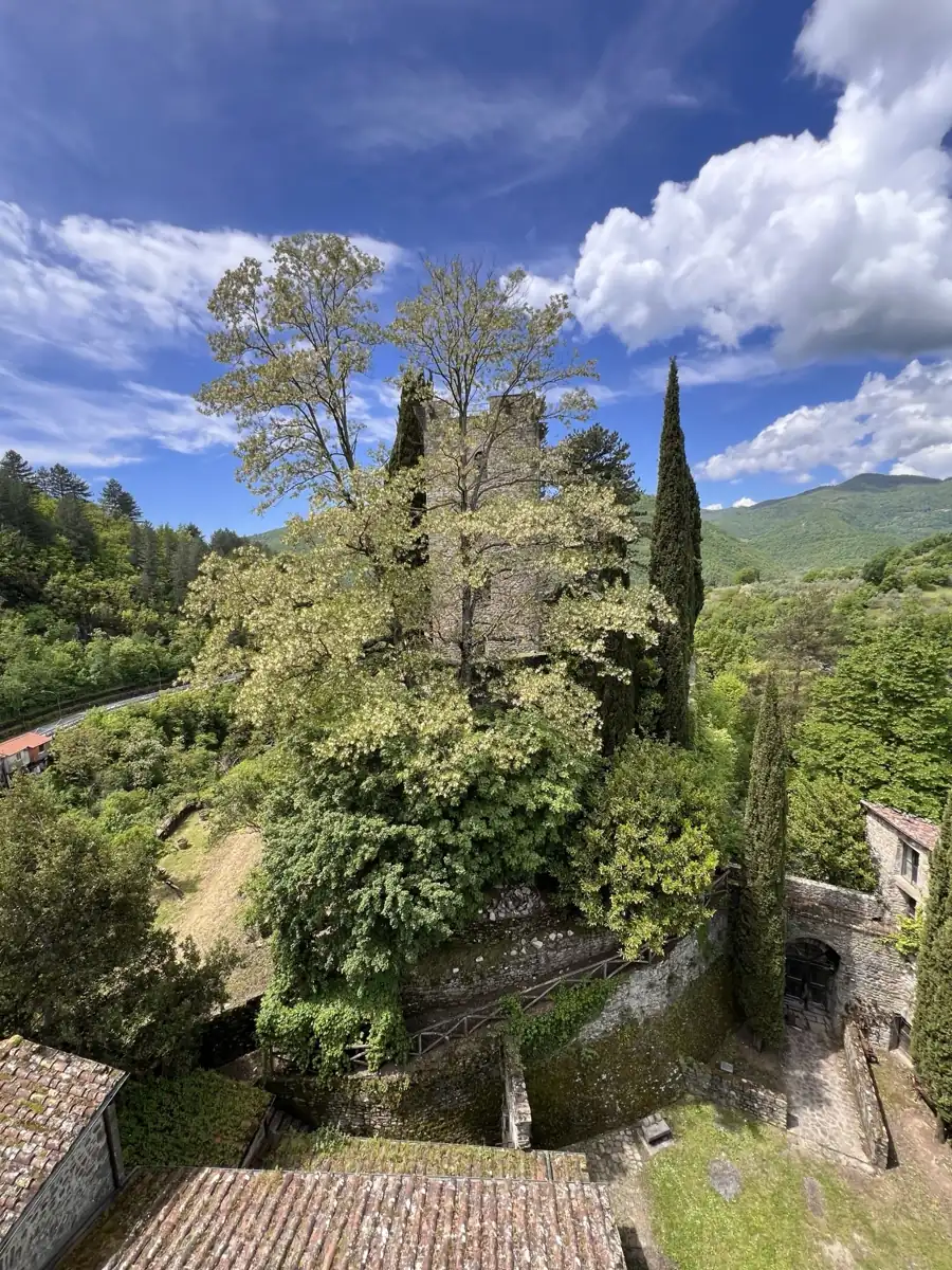 Il Castello della Verrucola a Fivizzano visto dall'alto: la torre tra alberi e cipressi, le mura in pietra e il cortile, sotto un cielo azzurro con nuvole e le colline della Lunigiana
